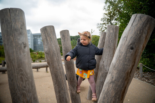 A little girl wearing a cat ear headband and black puffer jacket explores a wooden obstacle course at an eco-friendly playground in a park on a cold autumn day. Childhood curiosity, outdoor activity.