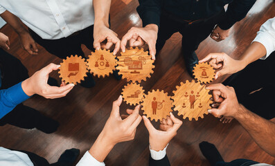 A diverse group of hands holds wooden gears featuring various business symbols, representing teamwork and collaboration in a modern professional environment. Amity