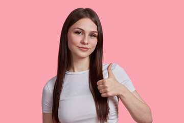 The girl shows the like sign with one hand. Portrait of a young beautiful student girl in a white T-shirt isolated on a pink background