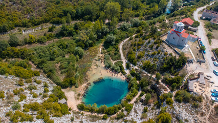 Aerial view of a vibrant blue lake nestled amid rugged terrain, contrasted by a stone church with a red roof, framed by lush green trees, Knin, Sibenik-Knin County, Croatia.