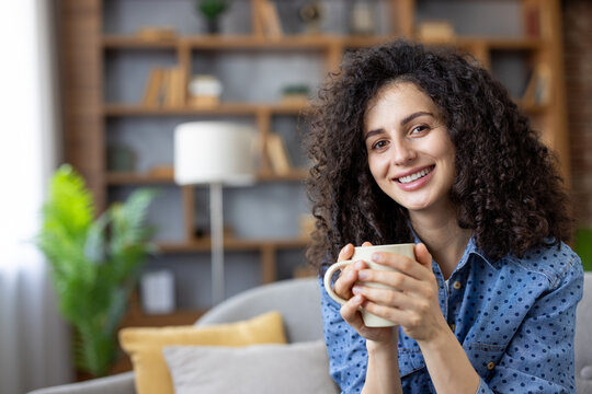 Smiling woman with curly brown hair relaxing on a cozy sofa in her modern living room, holding a warm mug and enjoying a peaceful morning break, content and comfortable