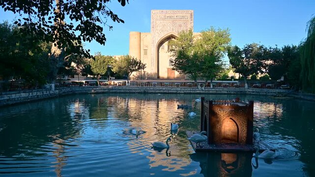 A calm daytime view of the central pond at Lyabi Hauz Square in Bukhara, Uzbekistan, surrounded by traditional buildings and shaded by trees.