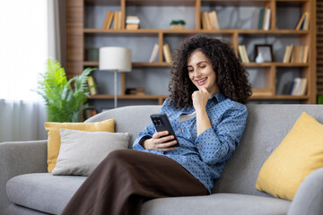 Young woman with curly hair relaxed on a gray sofa in a cozy living room, smiling as she scrolls her smartphone, enjoying social media, streaming and casual browsing