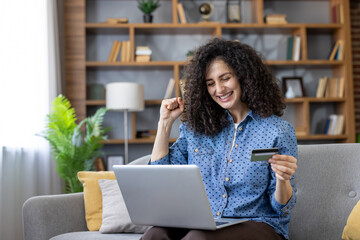 Young woman triumphantly purchasing goods online with a credit card, using a laptop, happily sitting on a couch in her living room, celebrating a successful transaction