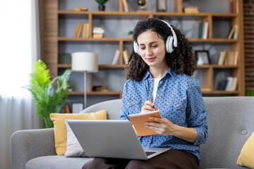 Young woman with curly hair wearing headphones and taking notes in a notebook, while sitting on a sofa and using a laptop for online learning or remote work at home