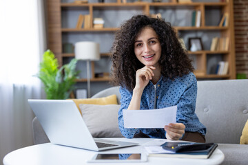 Woman with curly hair smiling and looking at camera while holding a paper document, sitting on a...