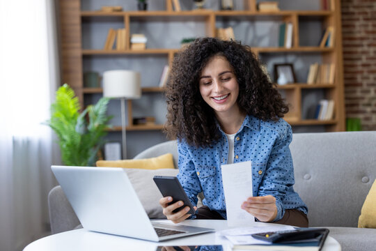 Woman sitting on a sofa, happily reviewing paperwork, holding a smartphone, and using a laptop to manage her budgeting, banking, and online bill payments at home