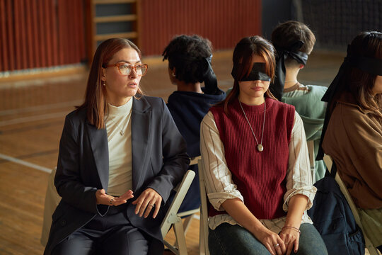 Caucasian young adult woman sitting beside blindfolded diverse group of students participating in psychology experiment