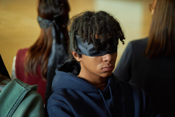 Teenage Black boy sitting with eyes covered by black blindfold participating in psychology experiment with other students visible from behind in classroom setting