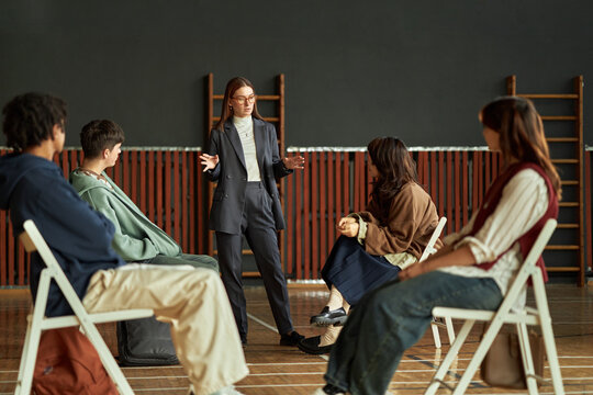 Caucasian young adult woman standing and gesturing while explaining psychological concept to diverse group of teenagers sitting on chairs in classroom setting during lesson