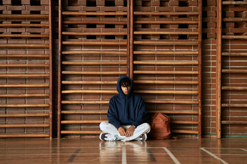 Black teenager boy sitting cross legged on gym floor in front of wooden wall bars wearing hoodie looking down with backpack beside him representing student psychology concept