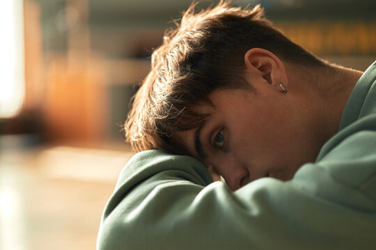 Portrait of Caucasian teenage boy resting head on arms, gazing thoughtfully into distance, showing contemplative expression, soft focus background suggesting educational environment