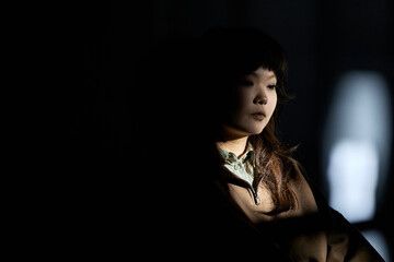 Asian young adult woman sitting in partial shadow, looking down thoughtfully, dark hair visible, appearing deep in contemplation, soft light highlighting face