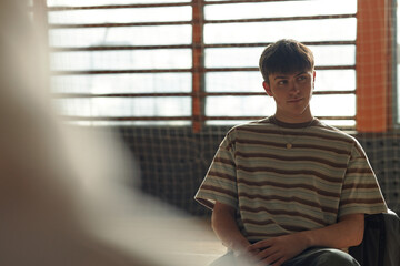 Caucasian teenage boy sitting in classroom setting listening attentively during psychology lesson, short brown hair, neutral expression, sunlight streaming through window blinds