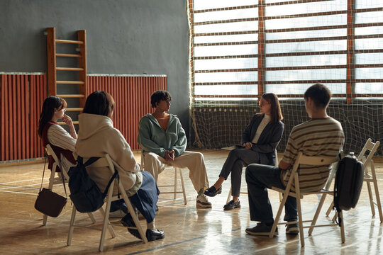 Group of multiethnic teenagers and young adult Caucasian woman sitting in circle discussing psychology topics in classroom setting, students engaging in group therapy or counseling session