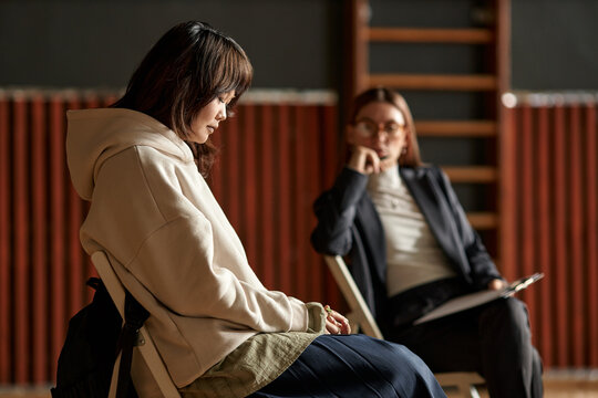 Asian female teenager sitting with head down during psychology session while Caucasian young adult woman psychologist observing and taking notes in educational setting