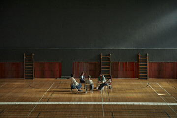 Group of multiethnic teenagers sitting in circle on chairs discussing psychology topics in gymnasium, engaging in collaborative learning activity as part of student group session