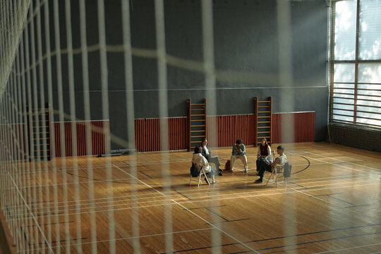 Group of multiethnic teenagers sitting in circle discussing psychology topics in sports hall, engaging in collaborative learning activity as sunlight streams through large windows