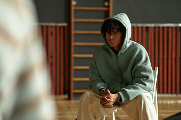 Black teenager boy sitting on chair with hands clasped, wearing hooded sweatshirt, looking forward with serious expression during psychology class or student counseling session
