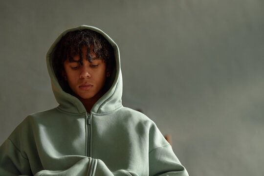 Teenager Black boy sitting with eyes closed wearing hooded sweatshirt, appearing thoughtful, psychology session concept, neutral background