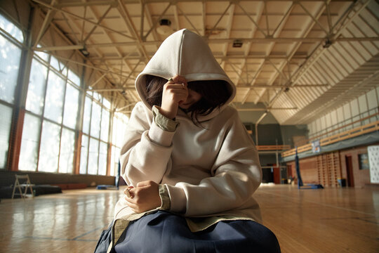 Teenage Asian girl sitting alone in gymnasium covering face with hand, wearing hooded sweatshirt, appearing withdrawn, adolescent emotional behavior concept