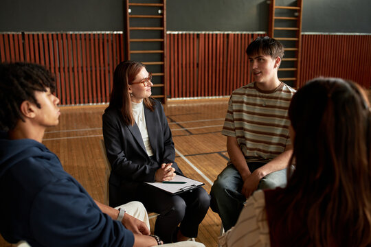 Group of multiethnic teenagers sitting in circle participating in psychology class with Caucasian woman instructor leading discussion in school gymnasium setting
