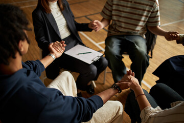 Group of multiethnic teenagers and young adults sitting in circle holding hands participating in psychology class session with clipboard on lap of Caucasian woman facilitator
