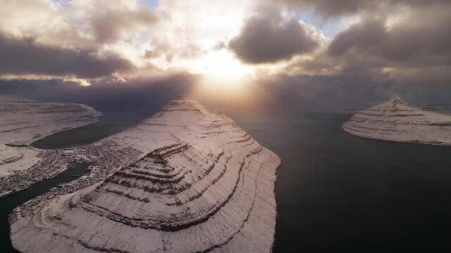 Aerial view of a snow-covered landscape with mountains, a town, and water under a cloudy sky, showcasing the contrasting white and dark tones, Streymoy, Streymoy, Faroe Islands.