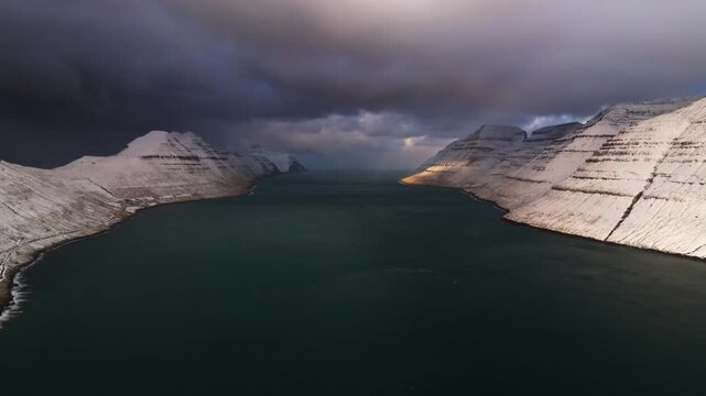 Aerial view of dramatic snow-capped mountains meeting the dark water under a moody sky, a serene contrast of light and shadow, Streymoy, Streymoy, Faroe Islands.