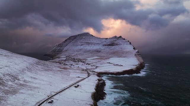 Aerial view of snow-dusted mountains meeting the dark, churning ocean under a dramatic sky, creating a stark contrast of textures and tones, Streymoy, Streymoy, Faroe Islands.