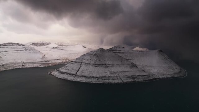 Aerial view of snow-dusted mountains rise from the dark waters, under a heavy, brooding sky, creating a stark contrast of light and shadow, Streymoy, Faroe Islands.