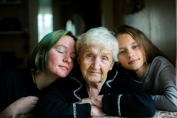 An elderly woman sits at a table with two granddaughters leaning close on each side, sharing a warm, intimate family moment in soft natural light.