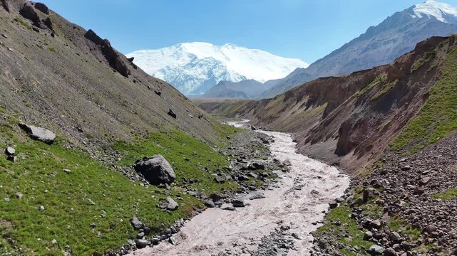 Drone footage captures a muddy river flowing through a dramatic mountain gorge, originating from the Alay Range. In the background, snow-covered peaks rise above the rugged terrain under clear skies.