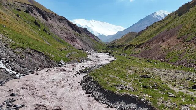 Drone footage captures a muddy river flowing through a dramatic mountain gorge, originating from the Alay Range. In the background, snow-covered peaks rise above the rugged terrain under clear skies.