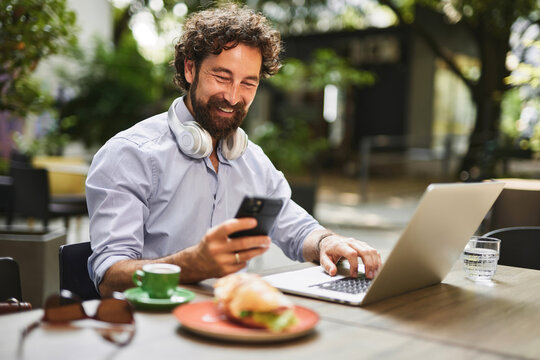 Man works on his laptop and checks his phone in a vibrant cafe surrounded by greenery, enjoying a breakfast of coffee and a sandwich during a sunny day.