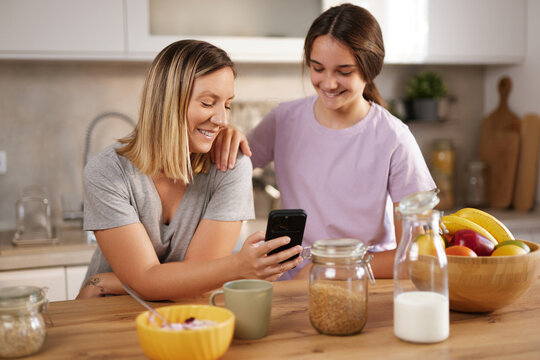 A mother and her daughter smile while looking at a smartphone in a bright kitchen, surrounded by fresh fruits and breakfast items, enjoying a relaxing morning together.