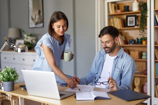 Two individuals are collaborating in a home office, with one reviewing documents while holding a coffee cup and the other writing notes at a desk with a laptop.