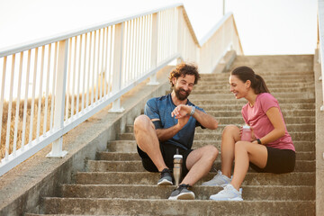 Two fitness enthusiasts relax on a stairway, sharing a light moment and checking their workout progress during a pleasant evening, enjoying a healthy lifestyle together.