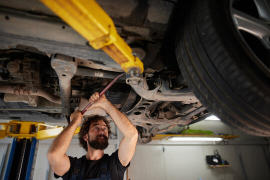 A skilled mechanic works on a car's undercarriage using a wrench while positioned under a hydraulic lift in a busy auto repair shop, ensuring quality maintenance and repairs.