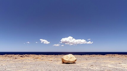 A single large rock rests on a desolate, rocky coastline with the deep blue ocean stretching to the horizon under a clear, bright sky.
