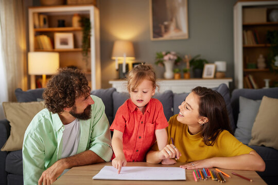 A family of three engages in a fun art activity at home. The child points to a drawing while the parents watch with smiles, creating a joyful atmosphere filled with creative expression.