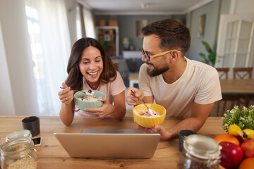 A couple shares a joyful moment over breakfast in their warm kitchen. They are eating from colorful bowls and looking at a laptop, smiling and engaging with each other's company.