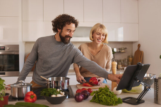 A couple enjoys cooking in a modern kitchen filled with fresh vegetables. The man is pointing at a device while the woman chops ingredients, creating a cheerful atmosphere. - Powered by Adobe