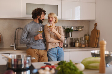 A couple stands together in a modern kitchen, laughing and raising glasses filled with sparkling...
