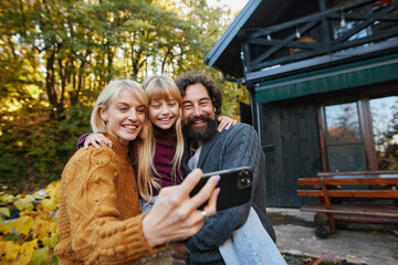 Three family members gather outside their cabin in autumn, smiling and taking a selfie. The trees...