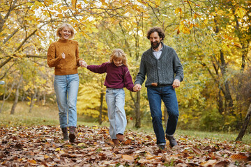 Naklejka premium A joyful family walks together on a sunny autumn weekend, surrounded by vibrant foliage and fallen leaves while sharing smiles and laughter in a park.