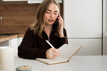 Woman student or freelancer multitasking in home office, phone call while taking notes in the kitchen