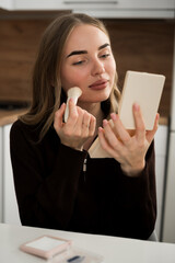 pretty woman applying blush on face holding small mirror sitting in home interior.