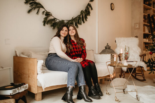 Happy mother and daughter sitting closely on a cozy sofa, embracing and smiling in a rustic home decorated with christmas greenery