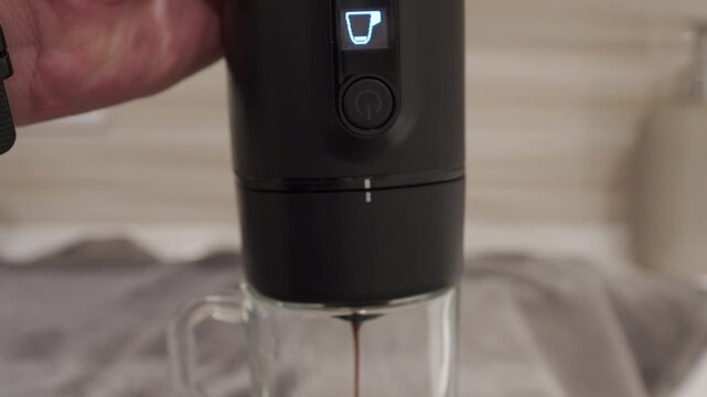 A hand holds a black plastic portable espresso machine as it brews and dispenses coffee into a glass mug. The internal electric pump makes a rattling and buzzing sound while it operates.